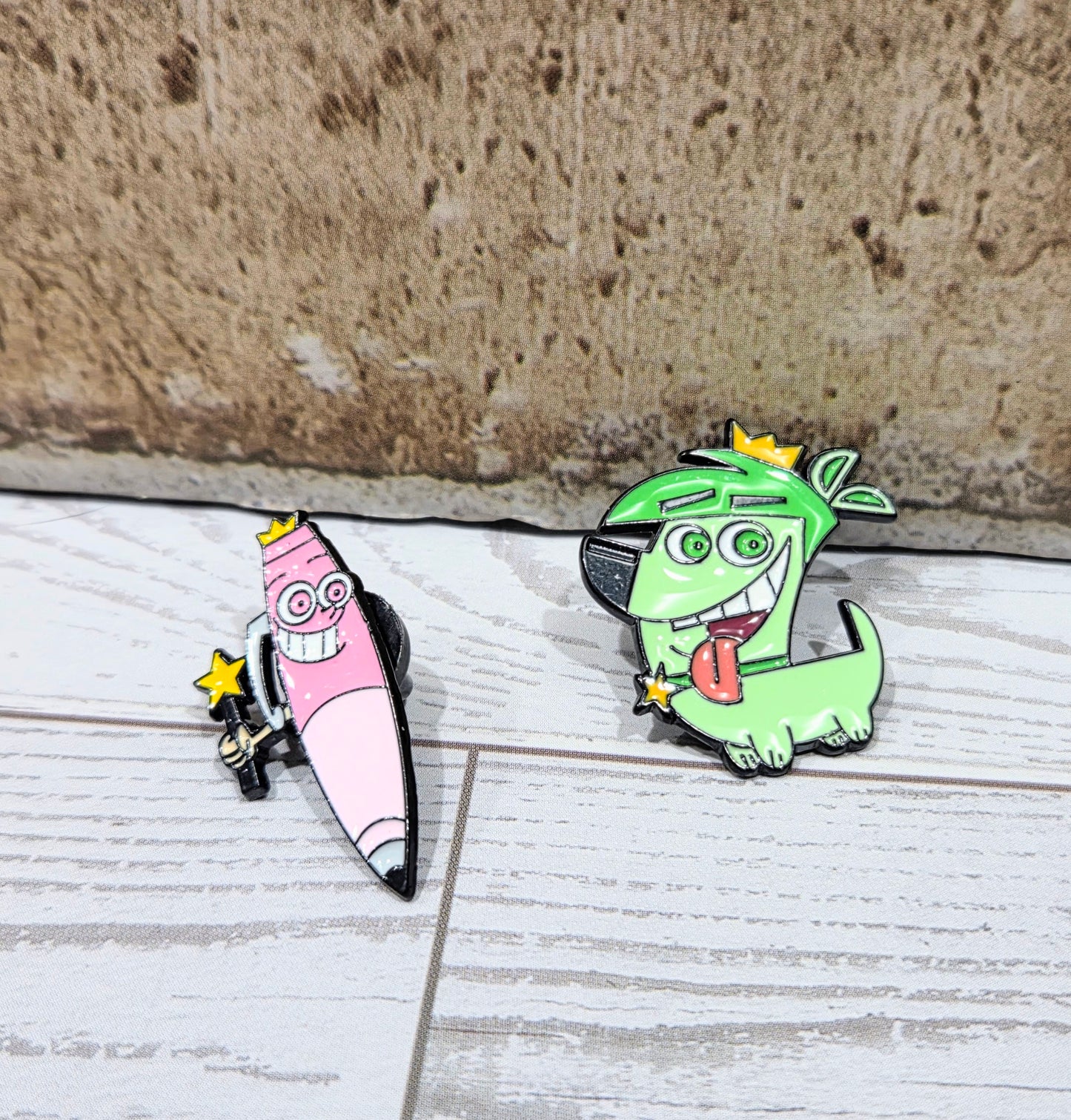 Two enamel character pins on a light surface: green and pink pencil designs