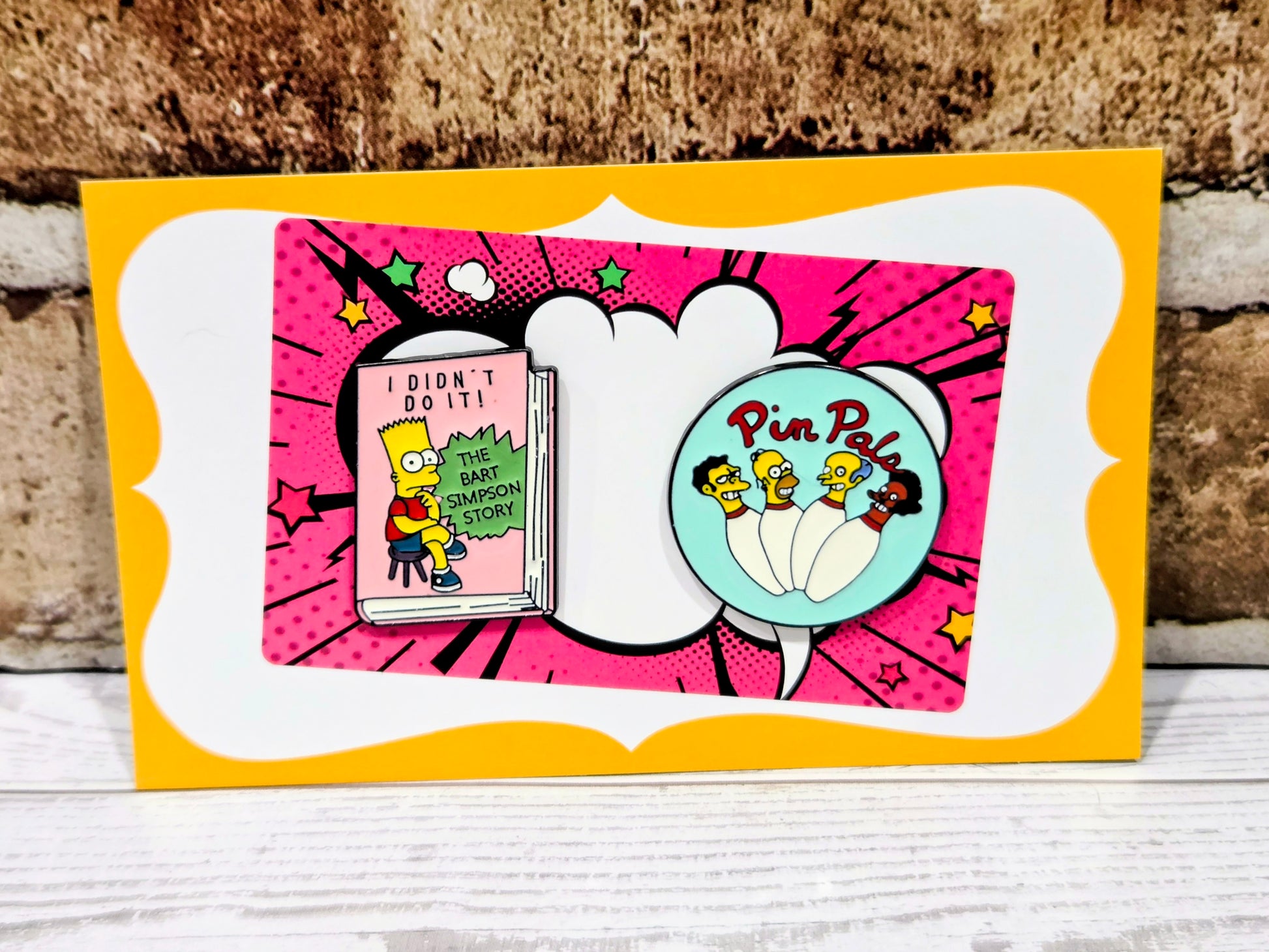Simpsons enamel pin set on retail card with round turquoise pin and pink book-shaped pin