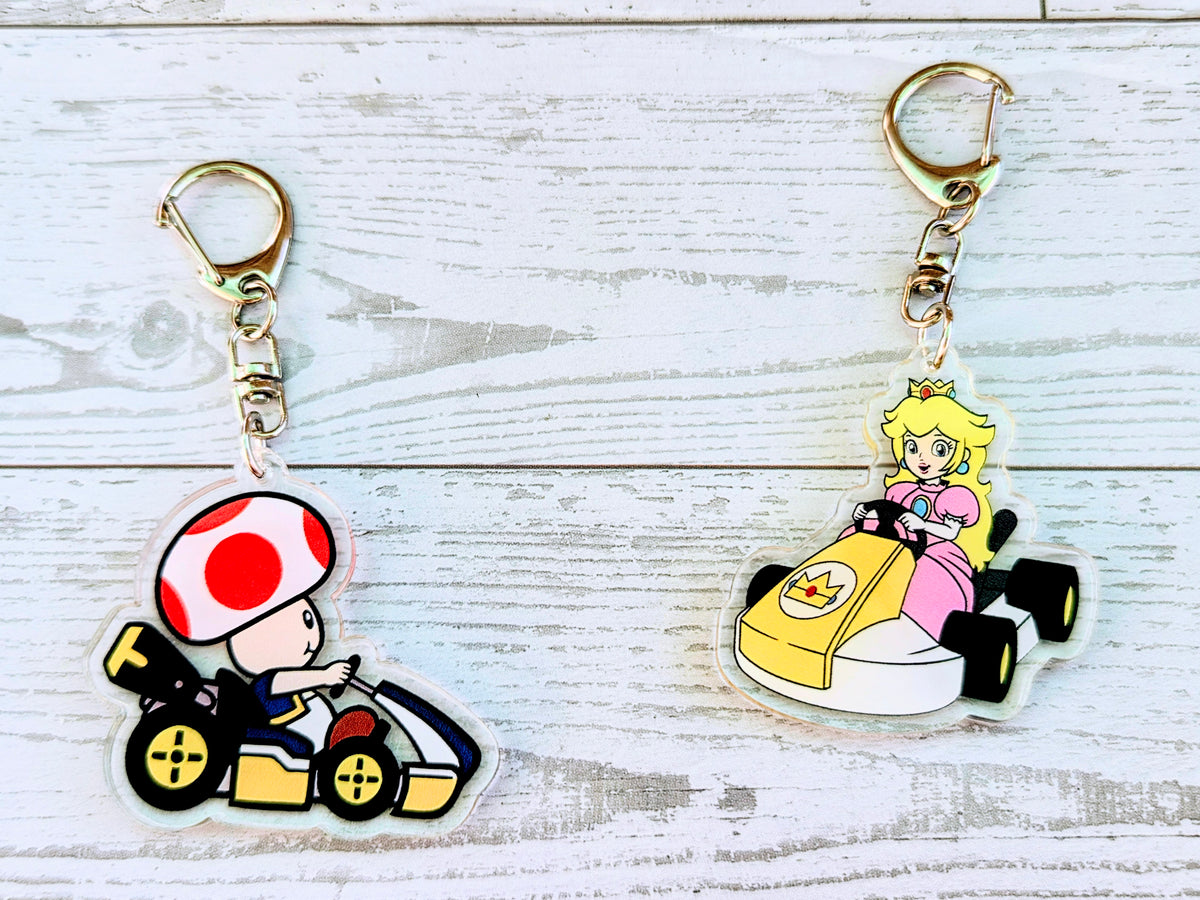 Two acrylic die-cut keychains: Toad in kart and Princess Peach in cart, gold keyrings