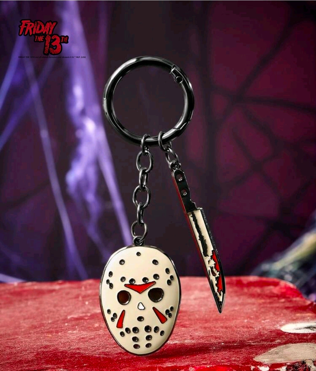 Metal keychain featuring oval mask enamel charm with red accents and a knife-shaped charm