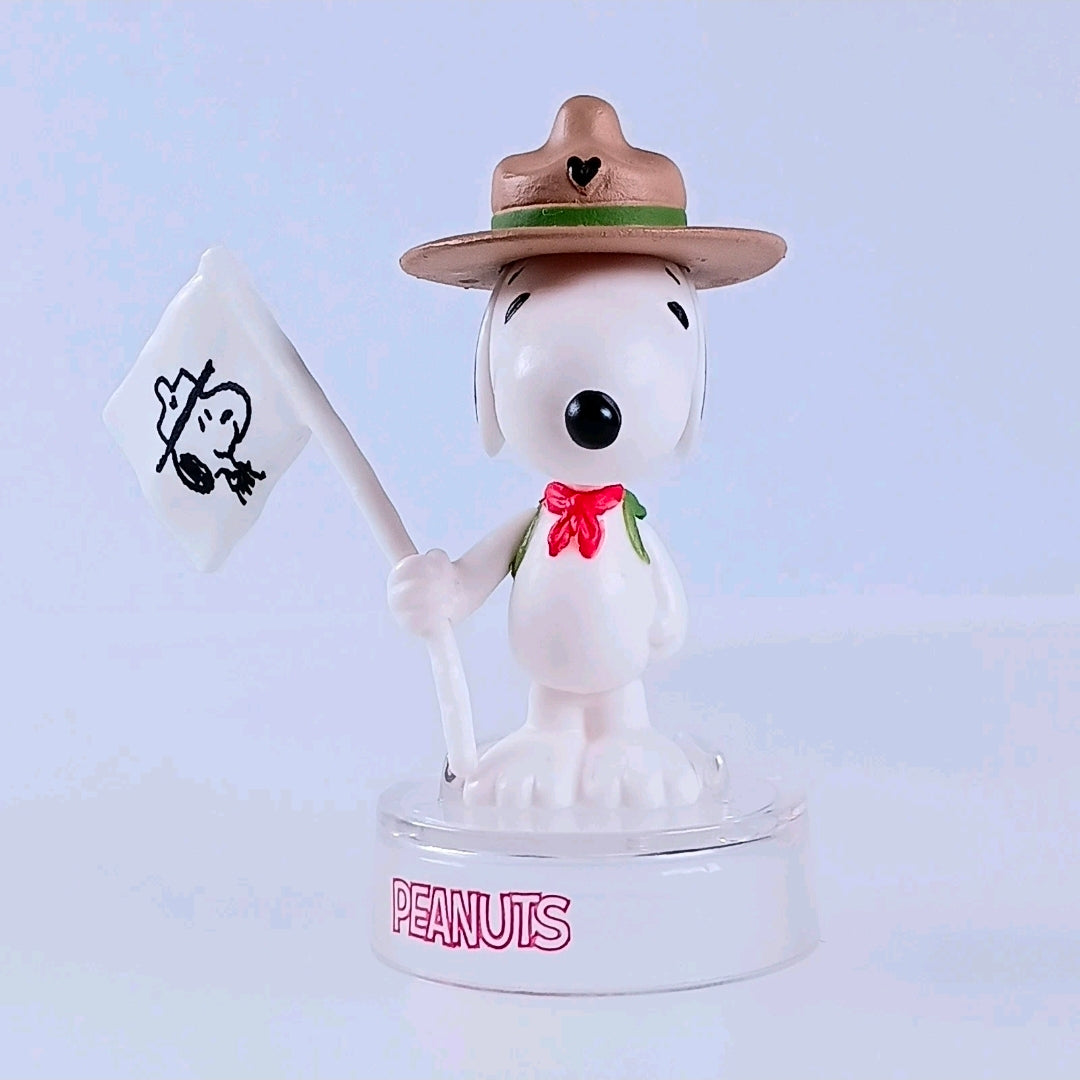 Snoopy figure in brown hat holding white sign, red ribbon bow, clear PEANUTS base