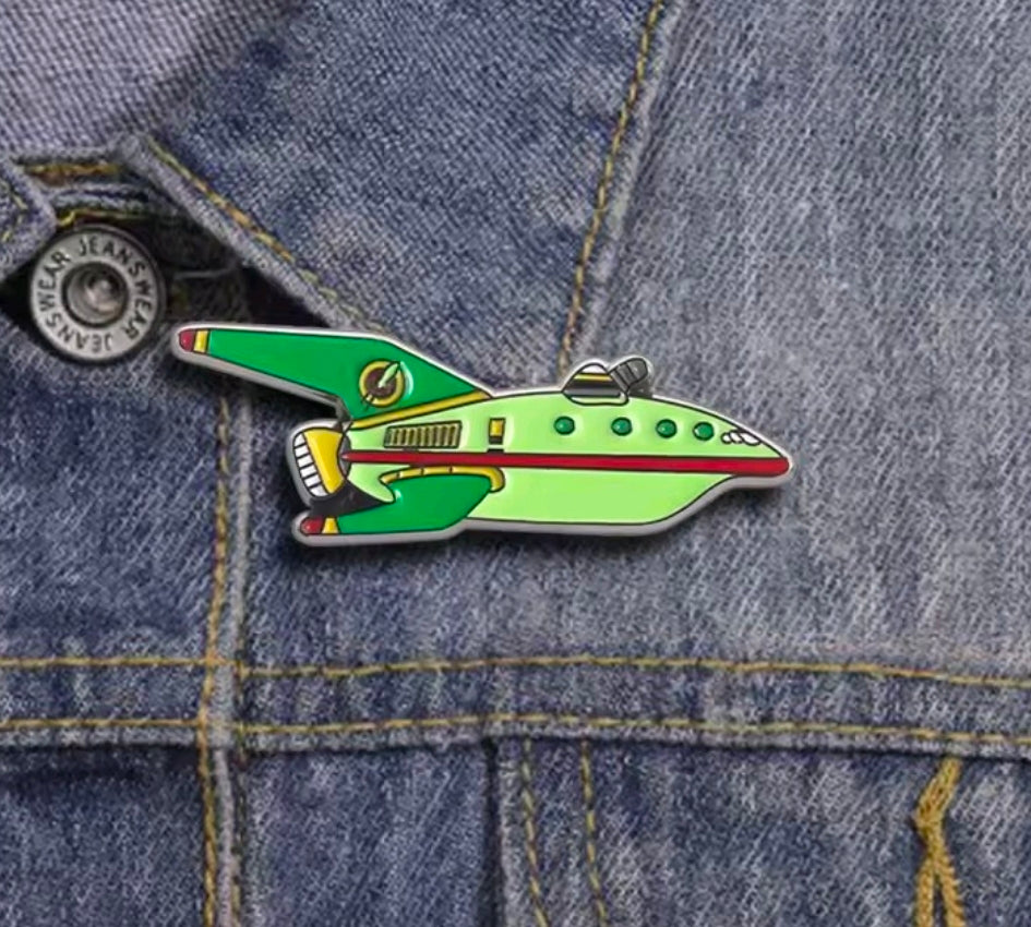 Light green spaceship enamel pin on blue denim with red and gold details