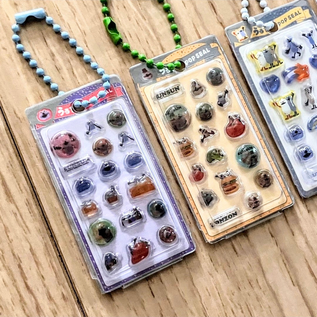 Assorted clear blister sticker keychains with beaded cords on a light wood tabletop
