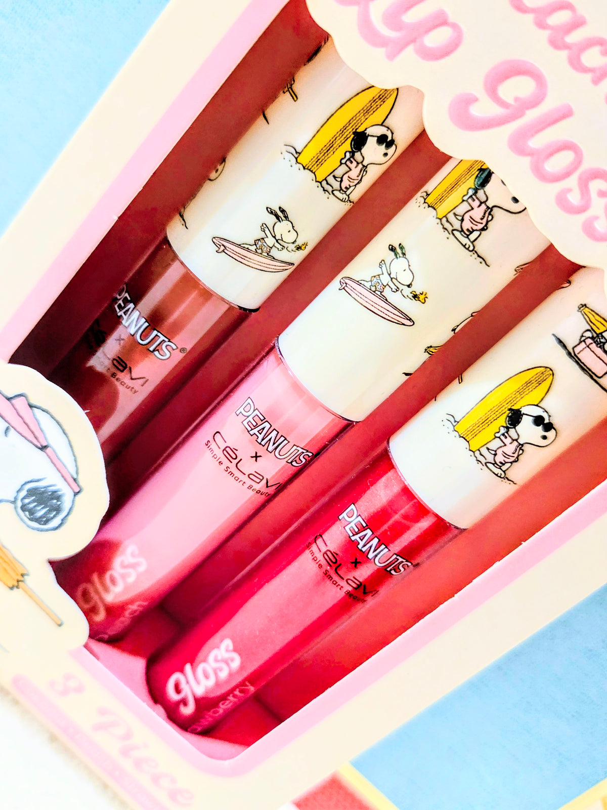 Three Snoopy lip gloss tubes in a pink tray, featuring beach-day character prints