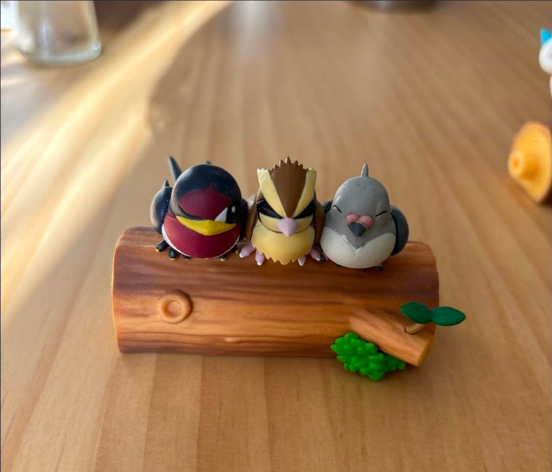 Three small bird-like toy figures on a brown log with scattered green sprouts.