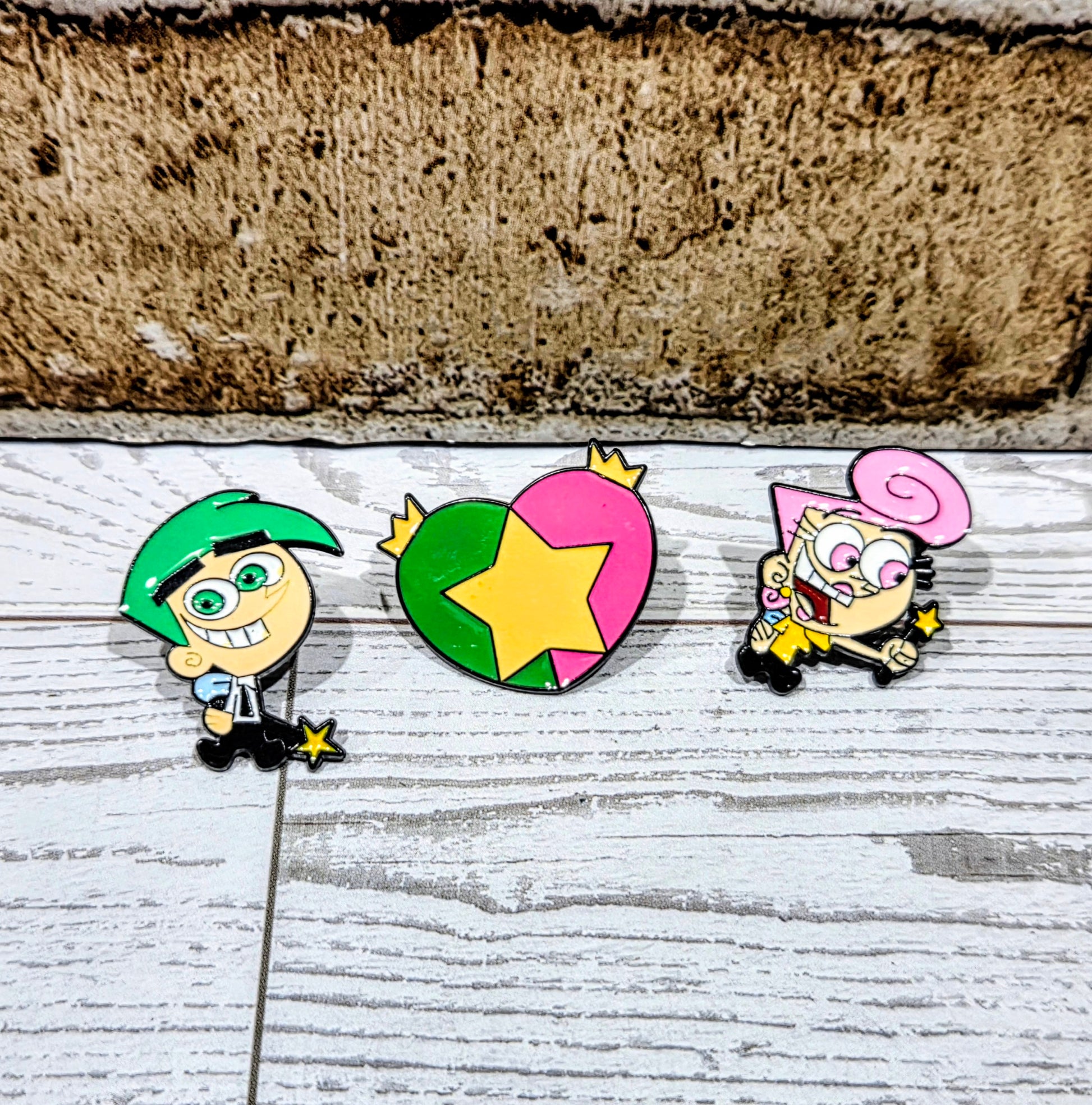Three enamel pins on display: Timmy character, green star-heart, and pink Wanda.