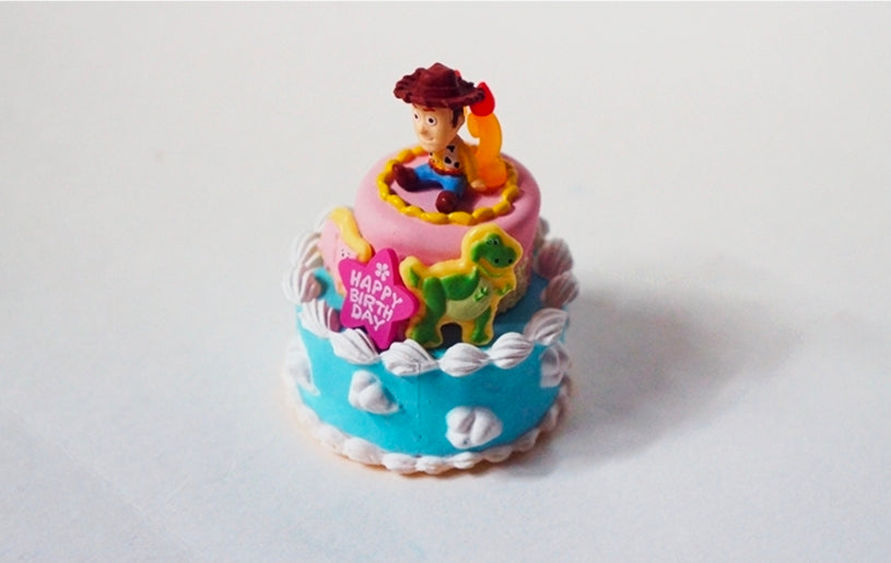 Mini birthday cake toy figure with Woody sitting on top, blue frosting and “Happy Birthday” sign
