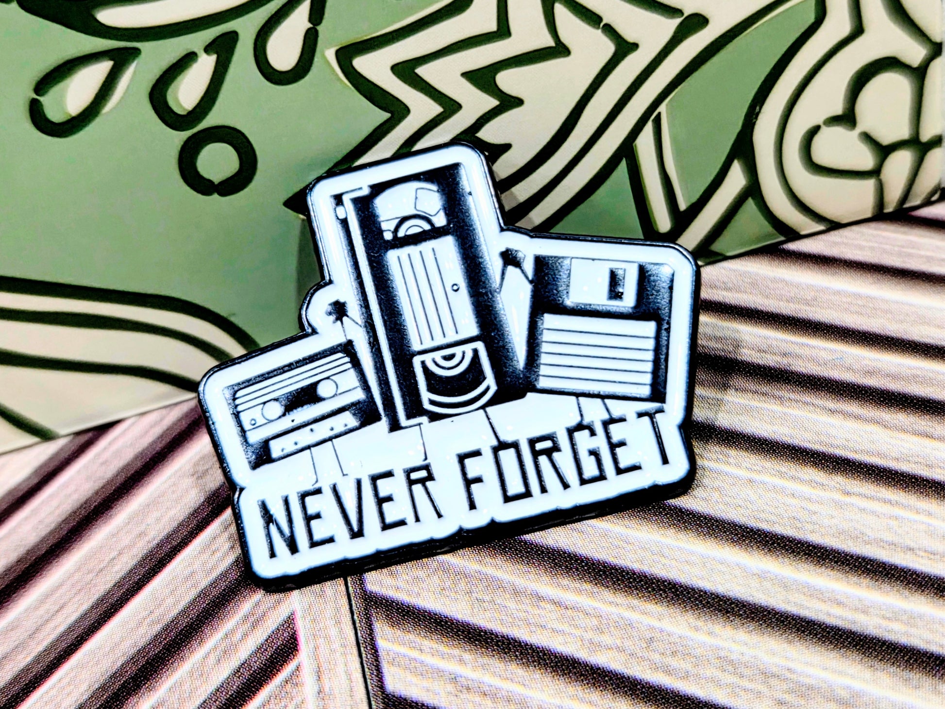Enamel pin shaped like a retro camcorder with text 'Never Forget'