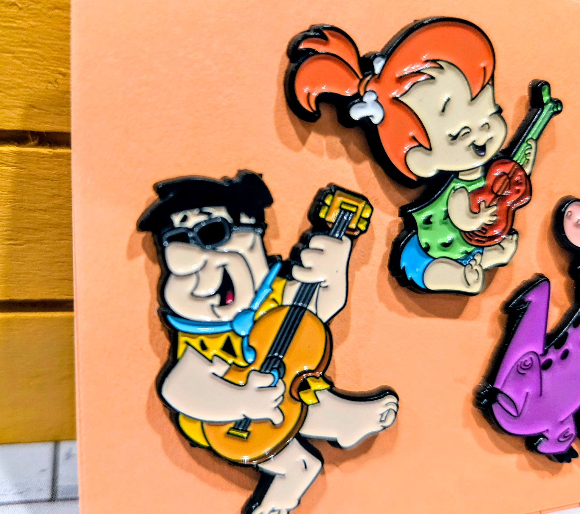 Enamel pin close-up: Fred, Pebbles, and Dino holding instruments