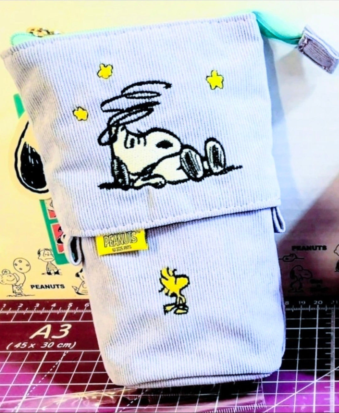 Lavender corduroy pop-up pencil case with Snoopy embroidery and PEANUTS tag, standing.