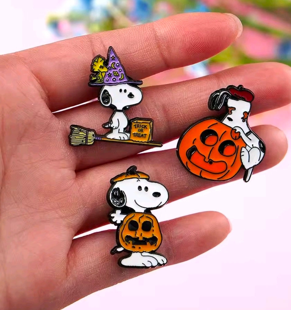 Hand holding Snoopy Halloween enamel pins: witch hat, trick-or-treat sign, and pumpkin.