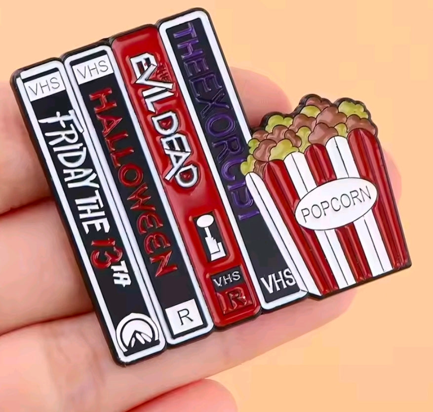 VHS tapes and popcorn enamel pin held in hand, red, purple, and black details.