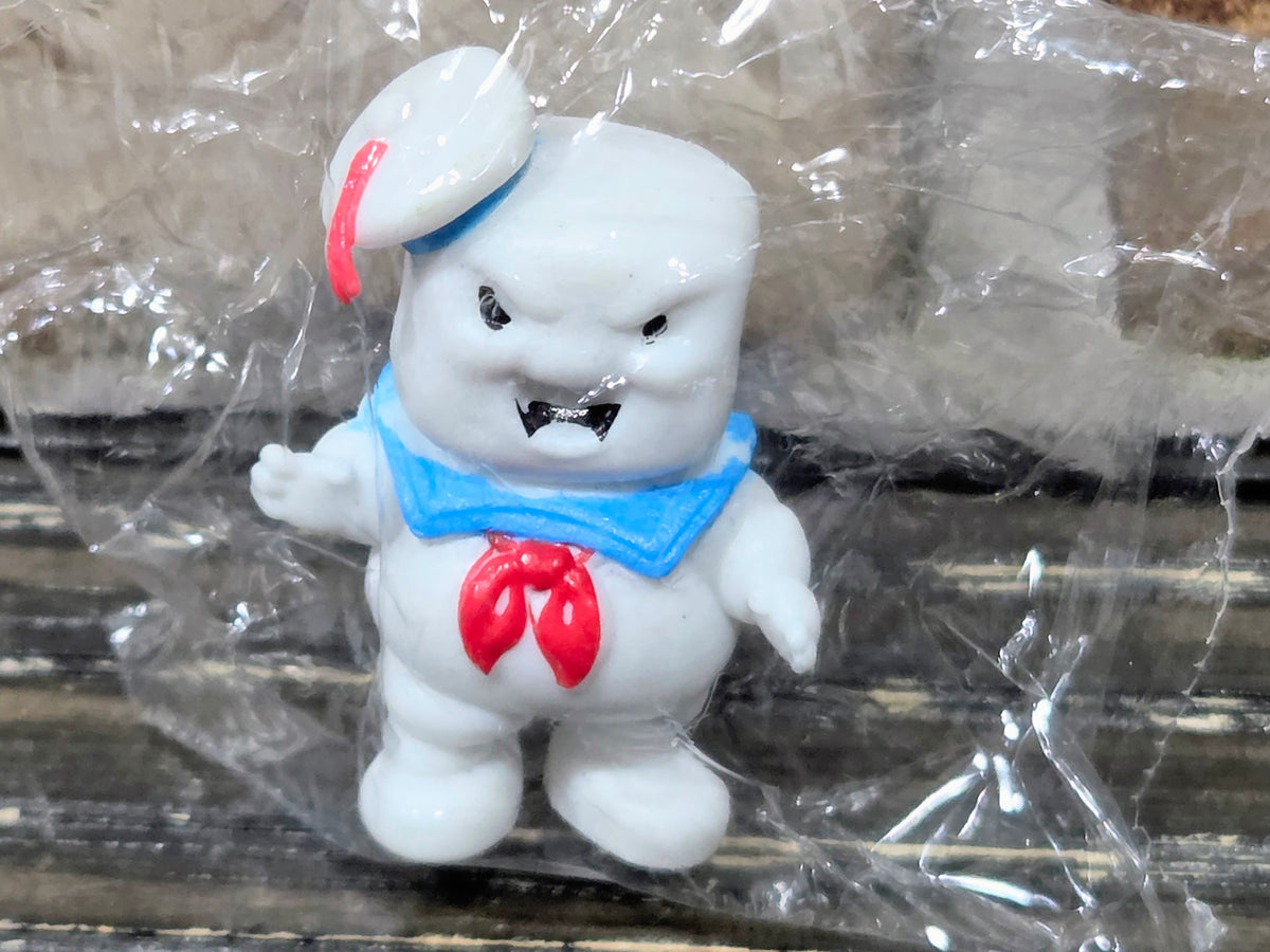 White mini figure standing, red hat, blue bib, red chest emblem, clear wrap