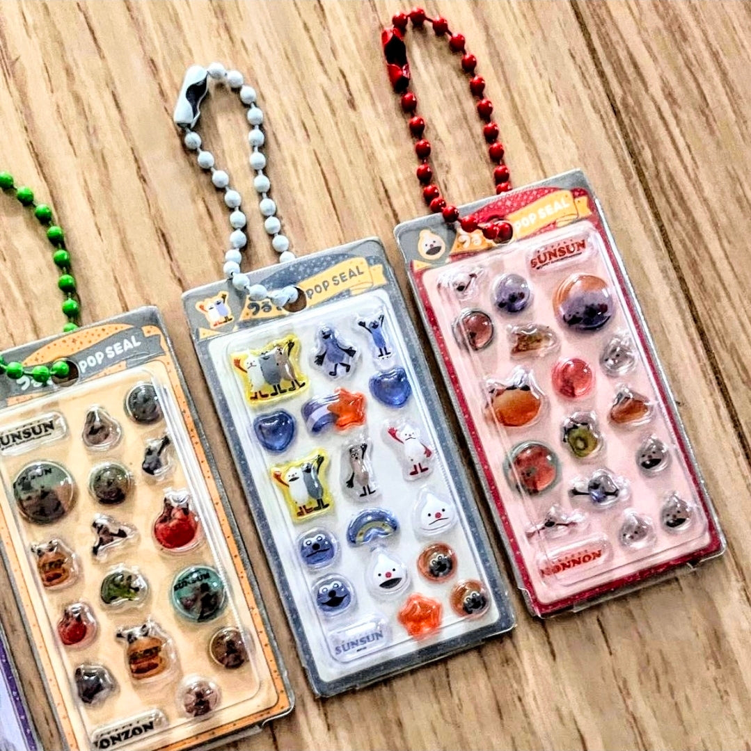 Three sticker keychain blister packs with beaded loops on a wooden surface