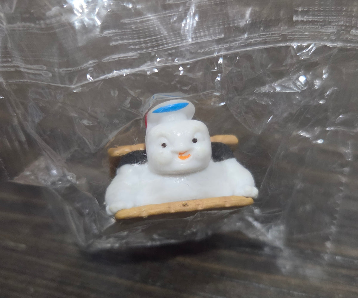 Toy mini figure with white marshmallow head on graham cracker base, sealed in clear plastic