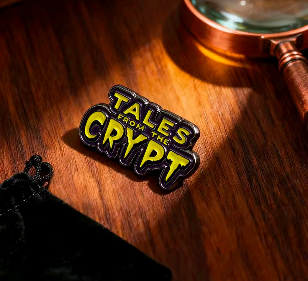 Tales from the Crypt enamel logo pin on wood, glossy black with yellow and purple text.