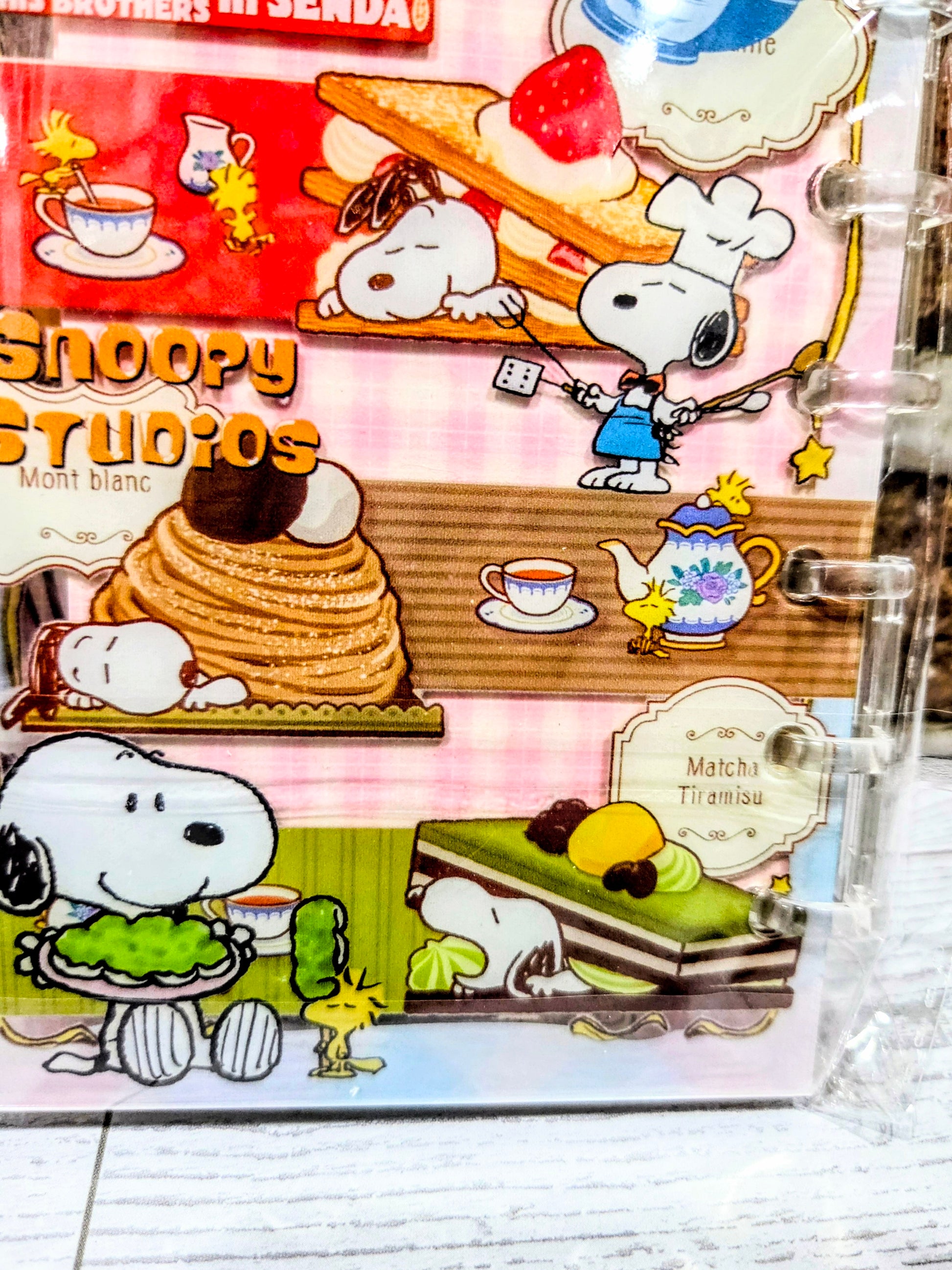 Pink acrylic shaker notebook close-up with Snoopy Studios cake and matcha tiramisu art