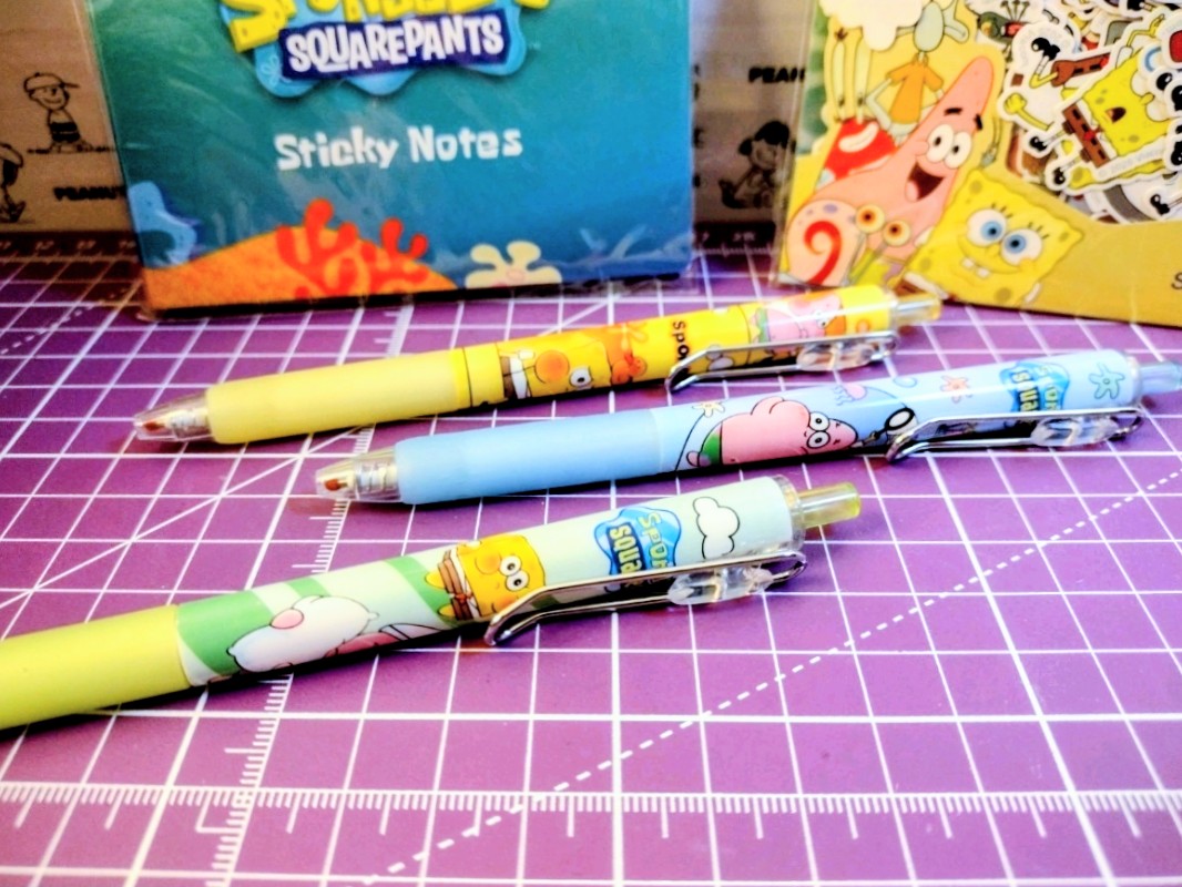 Three chibi character pens with colorful barrels and metal tips on a grid mat