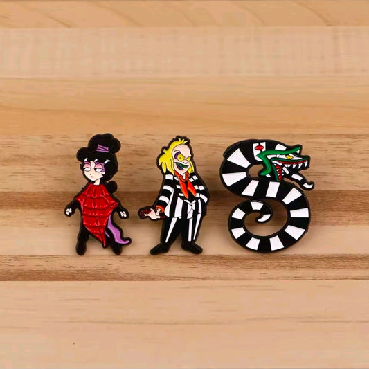 Three enamel character pins on wood: Lydia in red dress, Beetlejuice, sandworm