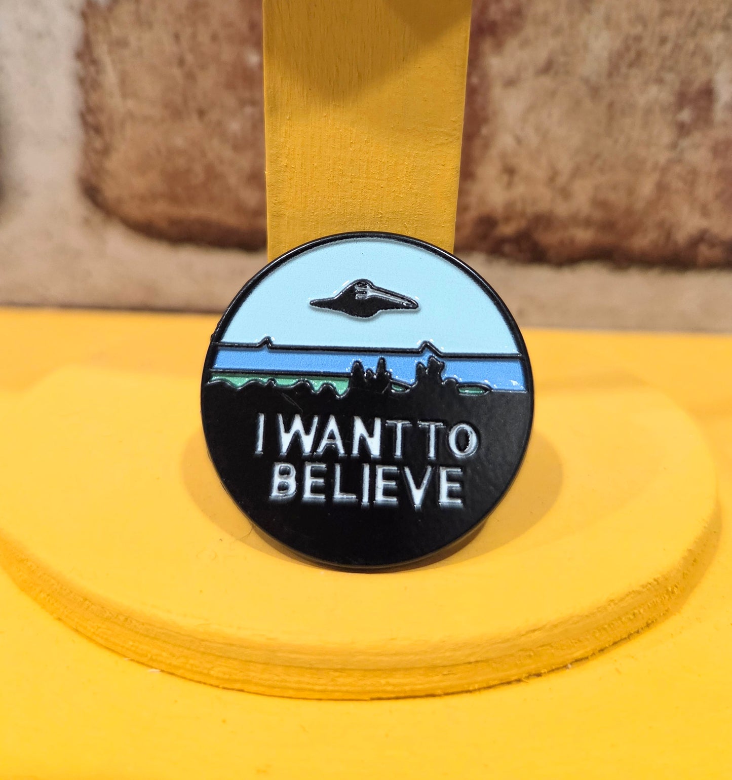 Round enamel pin with “I WANT TO BELIEVE” text, UFO silhouette, and blue landscape.