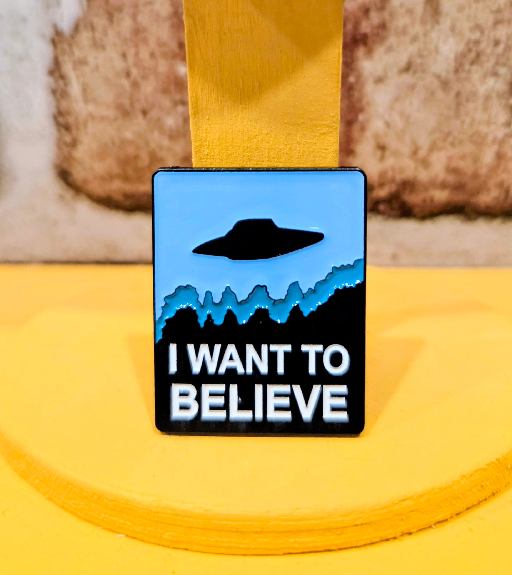 Square enamel pin featuring a black UFO and “I WANT TO BELIEVE” text on blue.