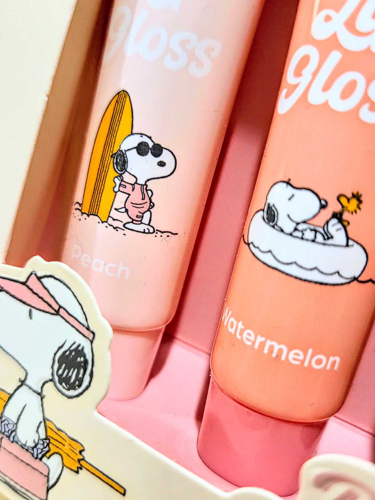 Close-up of peach and watermelon lip gloss tubes with Snoopy surfing illustrations
