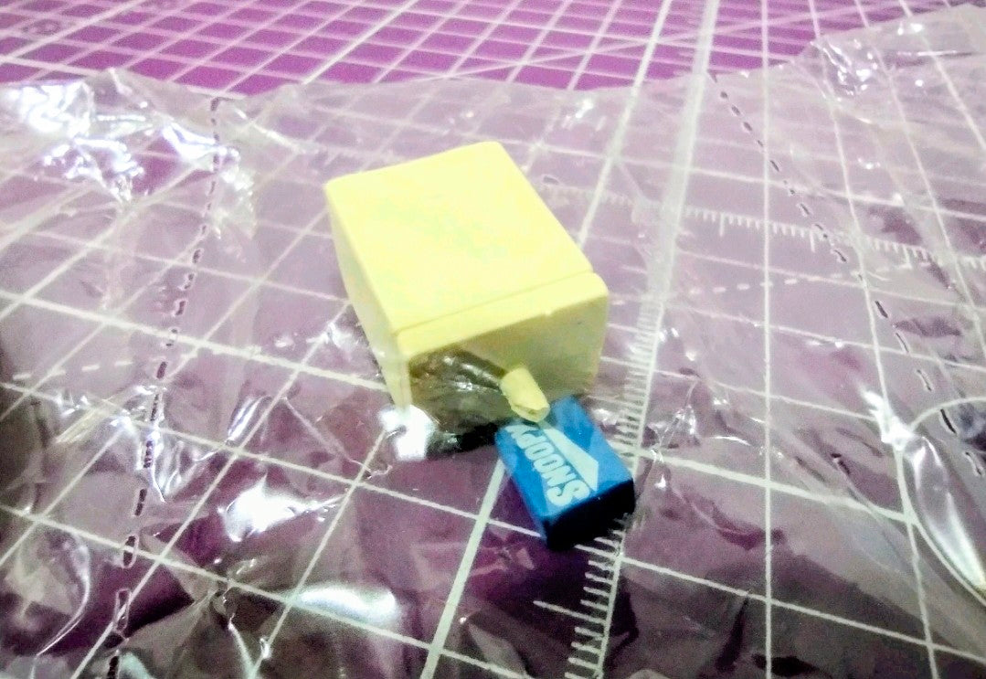Mini Snoopy accessory in blue with a small yellow block inside plastic wrapping
