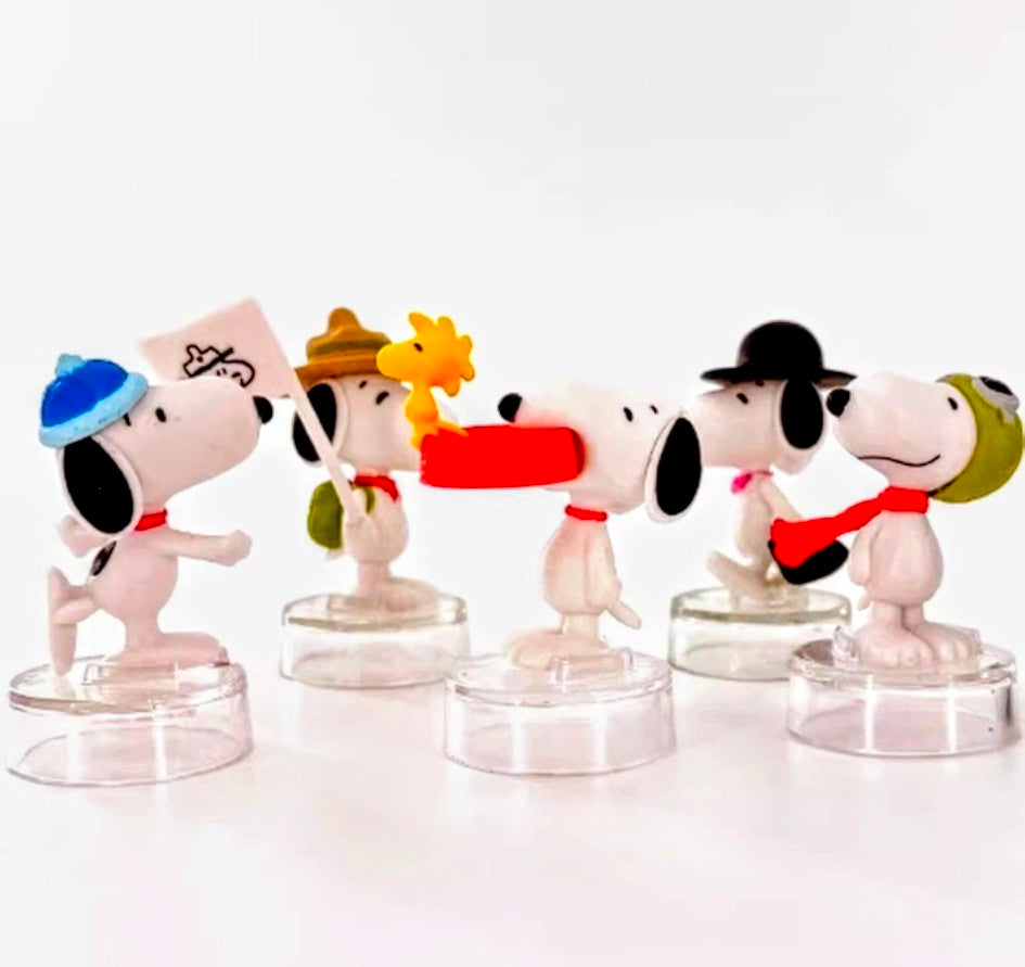 Five Snoopy figures lined up, wearing assorted hats and holding small props on clear bases