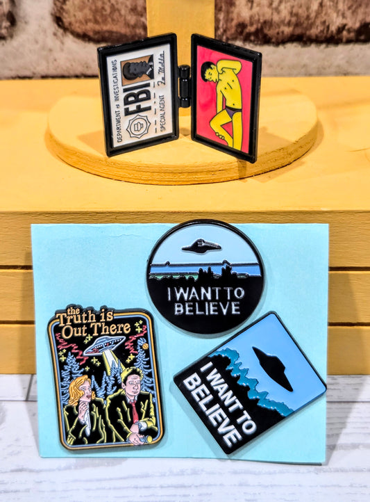 Enamel pin set on display stand, showing open hinged badges with alien and FBI designs.