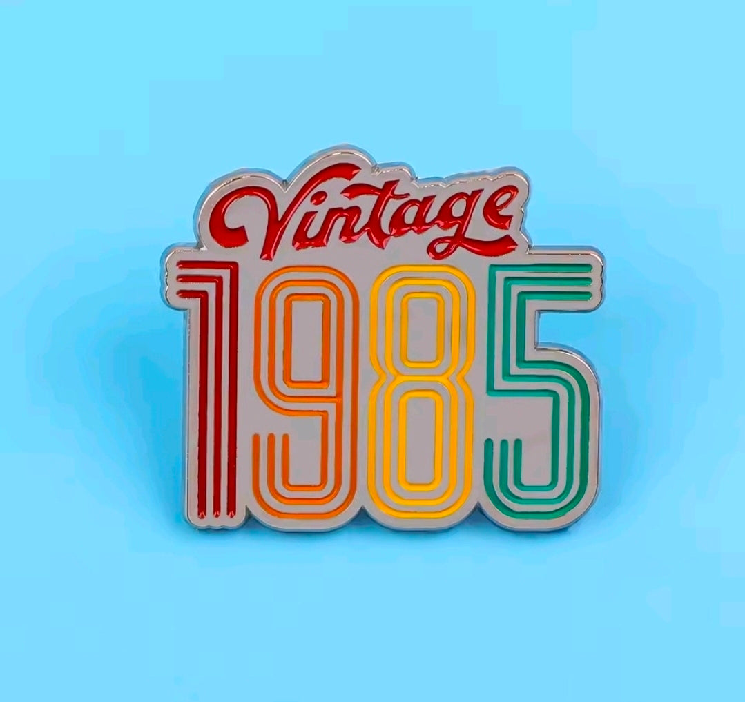 Enamel pin with 'Vintage 1985' rainbow striped lettering on display